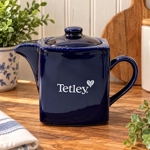 Könnte beinhalten: Eine quadratische, glänzend dunkelblaue Keramik-Teekanne mit passendem Deckel und geschwungenem Henkel. Auf der Vorderseite der Teekanne befindet sich der weiße Schriftzug Tetley mit einem kleinen Blattsymbol über dem Buchstaben y.
