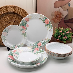 Puede incluir: Un conjunto de vajilla de cerámica blanca con un delicado patrón floral de rosas rosadas y hojas verdes. El set incluye platos y cuencos de varios tamaños con un diseño de borde sutilmente acanalado, dispuestos en pilas.