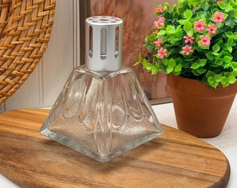 Könnte beinhalten: Eine Duftlampe aus klarem Glas mit quadratischem Boden und einer silberfarbenen, belüfteten Metallkappe. Der Glaskörper verfügt über einen texturierten, mattierten unteren Bereich und glatte, geschwungene vertikale Paneele zur Öffnung hin.