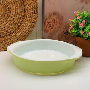May include: A round, shallow baking dish with a smooth, pale lime green exterior and a glossy white interior. The dish features two integrated, curved handles on opposite sides, providing a simple, functional design for kitchen use.