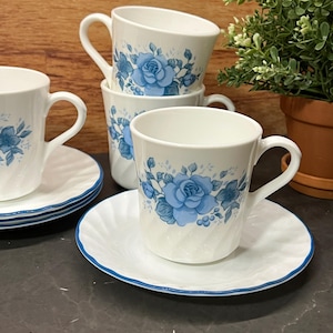 Könnte beinhalten: Weiße Teetassen und Untertassen mit blauem Rosenmuster. Die Tassen haben einen gebogenen Henkel und sind gestapelt. Die Untertassen haben einen geriffelten Rand mit blauem Rand. Eine kleine Topfpflanze befindet sich im Hintergrund.