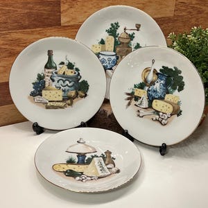 Peut inclure: Quatre assiettes en céramique blanche avec des bords dorés, chacune représentant une nature morte différente de fromage, de vin et d'ustensiles de cuisine. Les assiettes sont présentées sur des supports noirs sur un fond en bois.