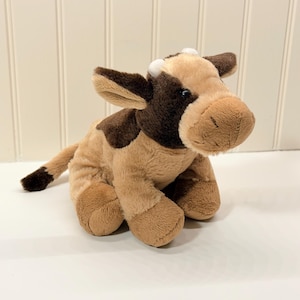 Peut inclure: Une peluche représentant une vache, aux tons marron et beige. L'animal en peluche présente des taches, des oreilles et l'extrémité de la queue marron foncé. Elle a de petites cornes blanches et est posée sur une surface blanche.
