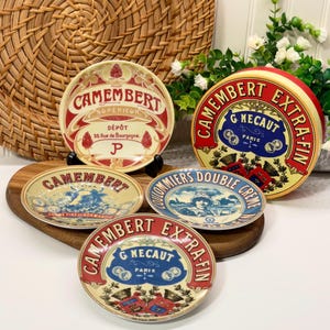 Peut inclure: Ensemble de plats à fromage Camembert vintage et une boîte à fromage ronde. Les assiettes présentent des motifs rouges, bleus et crème avec du texte français. La boîte ronde a une étiquette rouge et jaune. Les assiettes sont sur une planche en bois.