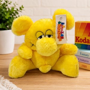 Peut inclure: Une peluche jaune vif avec de grandes oreilles, un museau arrondi et des yeux endormis. Une étiquette rectangulaire blanche est attachée à l'oreille, portant le texte Kodak Kolorkins avec des icônes de créatures colorées.