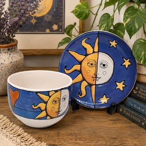 May include: A ceramic bowl and matching saucer set featuring a hand-painted celestial design. The bowl displays a sun and moon face with a heart, while the saucer shows a sun and moon face surrounded by yellow stars against a deep blue background.