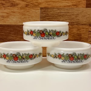 May include: Three white ceramic bowls with a decorative band of colorful vegetables and the text "Le Serveur". The bowls are stacked on top of each other, with the top bowl in focus. The background is a wooden surface.