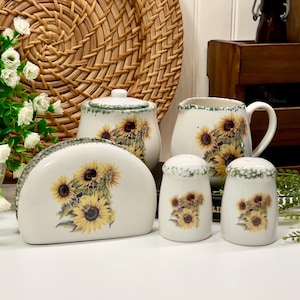 Peut inclure: Un ensemble de cuisine en céramique blanche avec un motif de tournesol. L'ensemble comprend un sucrier, une crémière, un porte-serviettes et des salières et poivrières. Chaque pièce a une bordure verte et une illustration détaillée de tournesol.