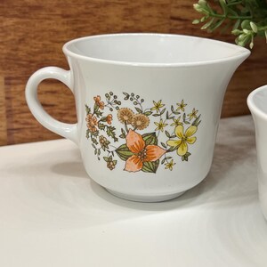May include: White ceramic creamer with a handle and a spout. The creamer is decorated with a floral pattern in orange, yellow, and brown. The creamer is on a white surface.