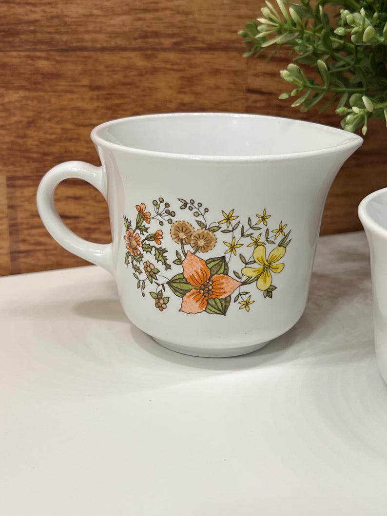 May include: White ceramic creamer with a handle and a spout. The creamer is decorated with a floral pattern in orange, yellow, and brown. The creamer is on a white surface.