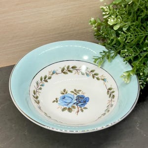 Może przedstawiać: Biała ceramiczna miska z niebieskim i zielonym wzorem kwiatowym. Miska ma jasnoniebieski brzeg z srebrną obwódką.