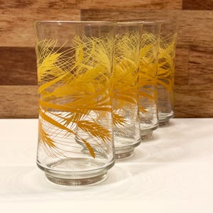 Peut inclure: Quatre verres à boire transparents avec un motif de tiges de blé jaunes. Les verres sont hauts et cylindriques, avec une base légèrement évasée. Le motif de blé est répété sur chaque verre, créant un ensemble cohérent. L'arrière-plan est un grain de bois clair.