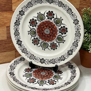 Peut inclure: Ensemble d'assiettes en céramique blanche avec un motif floral. Les assiettes présentent un motif floral central orange et marron entouré d'accents verts et noirs. Les bords sont décorés d'un motif floral noir et blanc.
