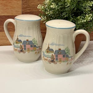May include: Two ceramic salt and pepper shakers with handles. The shakers are off-white with a blue rim and feature a painted village scene with houses, a church, and a horse-drawn carriage. The shakers are displayed on a white surface.
