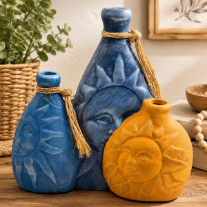 Puede incluir: Tres jarrones decorativos con relieves de caras de sol. Dos jarrones son azules con un acabado texturizado y uno es amarillo. Cada jarrón tiene un cuello estrecho atado con cuerda de yute natural. Los diseños muestran caras de sol en relieve.
