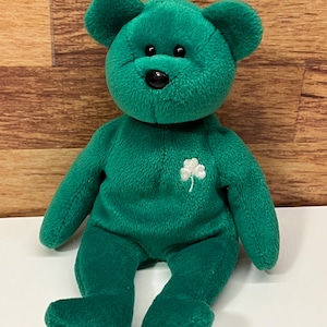 May include: A green plush bear with a black nose and small ears. The bear has a white shamrock embroidered on its chest. The bear is sitting with its legs extended. The background is a wooden surface.