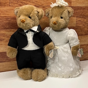 Peut inclure: Deux ours en peluche habillés pour un mariage. L'un porte un smoking noir avec une chemise blanche et un nœud papillon. L'autre porte une robe de mariée en dentelle blanche avec un coiffe assorti. Les deux ours ont des étiquettes.