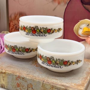 Peut inclure: Trois bols en verre opalin blanc ornés d'un motif végétal et de légumes colorés sur le pourtour. Chaque bol porte l'inscription Le Serveur en lettres cursives noires et présente un bord arrondi ainsi qu'une base légèrement conique.