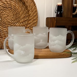 Peut inclure: Quatre mugs en verre dépoli avec des poignées, chacun avec un motif de nuage et de flocon de neige. Les mugs sont disposés sur une planche à découper en bois, avec un plateau en osier tressé en arrière-plan. Les mugs sont transparents avec un motif givré.