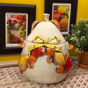 May include: A white ceramic pear-shaped cookie jar with a yellow bow and a lid decorated with colorful fruit, including strawberries, lemons, and oranges.