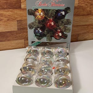 Puede incluir: Una caja de adornos navideños de cristal iridiscente. La caja presenta el texto "Festive Lights Boules Illuminées" y una representación de adornos en una rama de abeto. La caja contiene 12 adornos en forma de globo.