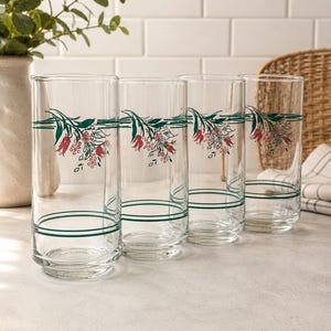 Pode incluir: Um conjunto de quatro copos de vidro transparente com um padrão decorativo de ramos florais e bagas em vermelho e verde. Cada copo apresenta duas linhas horizontais verde-escuras na base e uma linha perto da borda superior.
