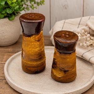 Peut inclure: Deux salières et poivrières en céramique avec un sommet brun foncé brillant et une base texturée jaune moutarde. Chaque récipient présente une forme cylindrique légèrement évasée au sommet avec quatre petits trous pour le dosage.