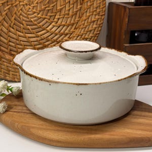 Puede incluir: Una fuente de cerámica blanca con tapa. El plato tiene una superficie moteada y un borde marrón. Se encuentra sobre una tabla de cortar de madera. La tapa tiene un pequeño asa. El plato está diseñado para cocinar y servir.