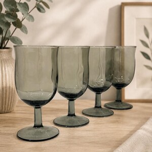 May include: A set of four smoke-gray glass goblets with a smooth, rounded bowl, a short stem, and a circular base. Each glass features a subtle, wavy texture along the sides, creating a uniform appearance across the collection of drinking vessels.