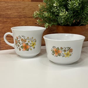 May include: A white ceramic teacup and bowl set. The cup has a handle and the bowl is round. Both pieces are decorated with a floral pattern in orange, yellow, and green.