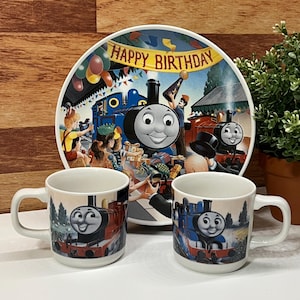 Puede incluir: Un juego de cumpleaños infantil con Thomas el Tren. El juego incluye un plato grande con el texto "Happy Birthday" y dos tazas. El plato y las tazas tienen ilustraciones coloridas de Thomas y otros personajes.