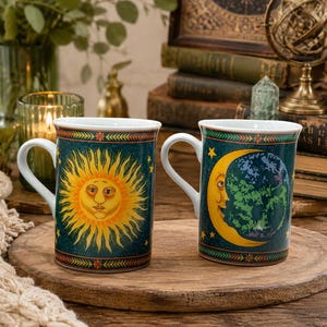 May include: Two ceramic mugs with white handles and interiors. One mug features a yellow sun with a face on a dark teal background with stars. The other mug displays a yellow crescent moon with a face and a blue and green earth on a dark teal background.