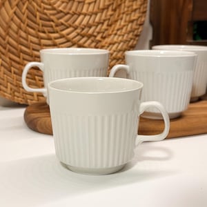 Peut inclure: Un ensemble de tasses en céramique blanche présentant une texture nervurée verticale sur l'extérieur. Chaque tasse possède une anse arrondie et lisse ainsi qu'un design épuré. Les tasses sont disposées en groupe, mettant en valeur leur forme uniforme.