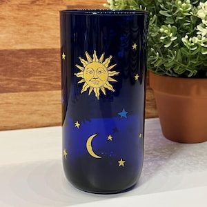 May include: A dark blue glass tumbler with a celestial design. The design includes a yellow sun, crescent moon, and stars. The glass is likely for drinking or decorative use, with a height of approximately 15 cm.