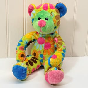 Peut inclure: Un ours en peluche coloré avec un motif floral vibrant dans les tons de vert, orange, rose et bleu. L'ours a un visage vert, un nez rose et des oreilles et des pieds bleus. L'ours est assis droit.