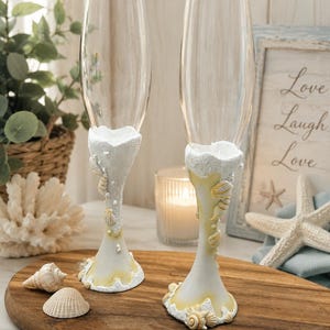 Peut inclure: Deux flûtes à champagne en verre transparent avec des pieds décorés de matière blanche texturée, de petits coquillages et d'accents perlés. La base de chaque pied présente une teinte jaune sable, évoquant un rivage de plage.