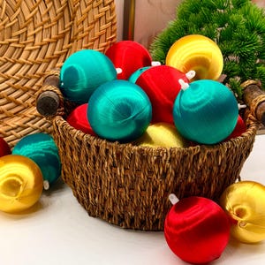 Peut inclure: Un panier en osier brun tressé rempli de multiples ornements ronds à la finition satinée dans des tons vifs de rouge, bleu sarcelle et jaune. Chaque ornement présente un petit capuchon en plastique blanc au sommet, et plusieurs sont éparpillés.