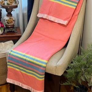 Peut inclure: Une couverture corail avec des rayures bleues, jaunes et vertes, drapée sur un fauteuil beige. La couverture est bordée d'une garniture en satin. Ce textile d'intérieur doux serait un ajout douillet à n'importe quelle pièce. Environ 150 cm de large.