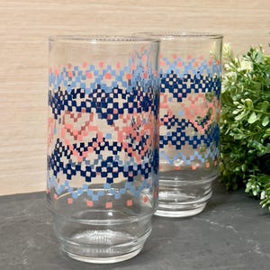 May include: Two clear glass drinking glasses with a blue, pink and white geometric pattern. The glasses are on a wooden surface.