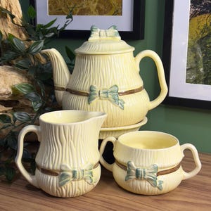May include: A yellow ceramic teapot, creamer, and sugar bowl set with a woodgrain texture and green bow accents.