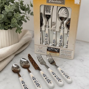 Peut inclure: Un ensemble de couverts Corelle Charlotte de 20 pièces en acier inoxydable pour quatre personnes, avec des manches blancs ornés d'un motif floral bleu et doré. Le set comprend des cuillères, des fourchettes et des couteaux dans une boîte en carton.