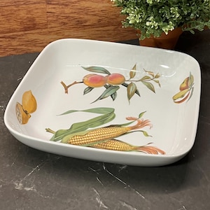 Peut inclure: Plat de service carré blanc avec un motif décoratif de fruits et de légumes. Le motif comprend des pêches, du maïs et un citron. Le plat a un bord légèrement incurvé et est posé sur un fond sombre.