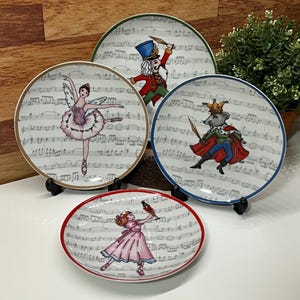 May include: Four decorative plates featuring ballet-themed illustrations. Each plate has a white background with musical notes and a colorful border. The plates depict characters from the Nutcracker ballet, including a ballerina, a soldier, and the Mouse King.
