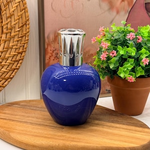 Puede incluir: Una lámpara de fragancia de vidrio en forma de corazón, de color azul intenso y acabado brillante, con una tapa de corona de metal plateado pulido. El recipiente tiene una silueta suave y redondeada con una superficie reflectante que capta la luz.