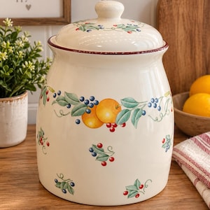 Peut inclure: Un pot en céramique de couleur crème avec un couvercle assorti, orné d'un motif peint d'oranges, de feuilles vertes et de petites baies bleues et rouges. Le pot a une forme cylindrique avec un bord légèrement évasé et une bordure rouge foncé.