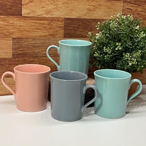 Peut inclure: Quatre tasses en céramique de couleurs pastel : rose, gris et deux nuances de vert d'eau. Chaque tasse a une anse incurvée. Une petite plante en pot est visible en arrière-plan. Les tasses sont disposées sur une surface blanche.