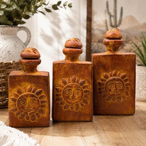 Peut inclure: Trois bouteilles rectangulaires de couleur terre cuite de hauteurs variables, chacune présentant un motif de visage solaire en relief sur le devant et un petit bouchon arrondi sur le dessus. Finition texturée et patinée orange et marron.