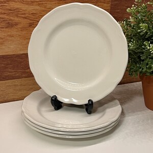 Puede incluir: Cuatro platos de cerámica blanquecina con bordes festoneados. Un plato se exhibe en un soporte negro. A la derecha hay una pequeña planta en maceta. Los platos tienen un diseño sencillo y elegante.
