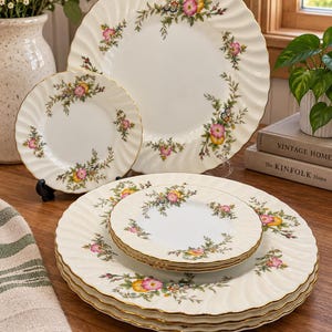 Peut inclure: Un ensemble d'assiettes en céramique couleur crème avec un bord festonné à liseré doré et un motif floral composé de fleurs roses, jaunes et bleues avec des feuilles vertes. La collection comprend de grandes assiettes et des petites assiettes.