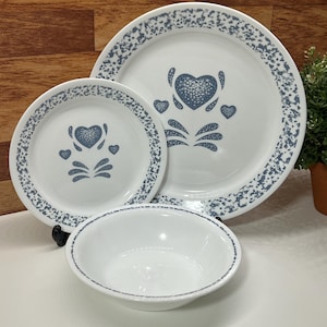 May include: A white dinnerware set with a blue heart design. The set includes a large plate, a smaller plate, and a bowl. The plates have a blue speckled border. The heart design is in the centre of the plates.